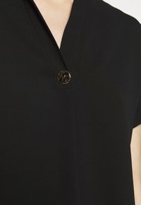 Haut noir à manches courtes avec une finition texturée et un décolleté en V, doté d'un bouton rond brillant unique comme détail distinctif.