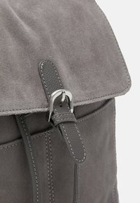 Szary zamszowy plecak z klapką, z metalowym zapięciem w kolorze srebrnym i ciemną skórzaną rączką. Widoczne szwy i gładka tekstura.