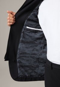 Wnętrze czarnej kurtki z szarym abstrakcyjnym wzorem, zapięcie na zamek oraz mała biała kieszonka akcentująca; gładka faktura materiału.