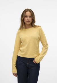 Gul strikket sweater med lange ærmer, rund udskæring og ribbede manchetter. Parret med mørkeblå jeans, har den en blød tekstur.