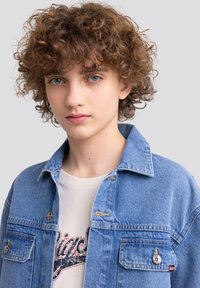 Giacca di jeans azzurro chiaro con colletto classico, tasche con bottoni e leggere usure, indossata sopra una t-shirt beige con stampa grafica.