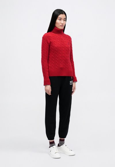 Pulover roșu cu guler înalt din tricot cu model, cu tiv elastic, purtat cu pantaloni de trening negri și adidași albi. Textură netedă, design ajustat și stil casual.