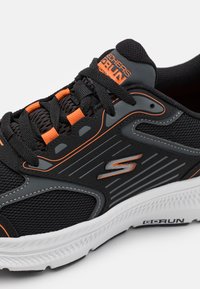 Zapatilla deportiva negra con parte superior de malla, acentos naranjas y detalles grises. Presenta una suela blanca con dibujo texturizado y un logo en el lateral.
