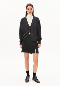 Frau trägt eine dunkelgraue, knöpfbare Strickjacke über einem weißen Rollkragenpullover, einen schwarzen Minirock mit seitlichem Schlitz, schwarze Socken und schwarze Loafers mit Nieten.