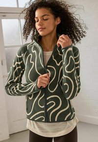 Groene fleece jas met een spiraalpatroon in crèmekleur, voorzien van een hoge kraag, ritssluiting en lange mouwen. Gelaagd over een lichte top.