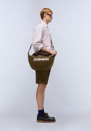 Olivengrøn skuldertaske med logo, brun rem, båret af person i stribet skjorte, shorts, marineblå sokker og sorte sko, set fra siden.