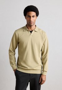 Világos olívazöld, hosszú ujjú, V-nyakú galléros póló mandzsettákkal. Sima anyagból készült, klasszikus gallérral és laza szabással.