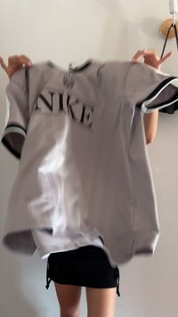 Camisola cinza da Nike com detalhes e logotipo em preto, mangas curtas, tecido leve, ligeiramente oversized, exibida contra um fundo neutro.