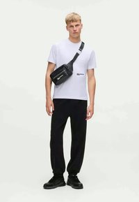 Zwarte crossbody tas met een gladde textuur, gedragen over een eenvoudige witte t-shirt en losse zwarte broek. Stevige zwarte sneakers maken de look compleet.