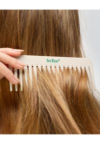 Biały plastikowy grzebień z szerokimi zębami, oznaczony "So Eco", trzymany w pobliżu długich, falujących brązowych włosów. Skupienie na wzorze grzebienia i strukturze włosów.