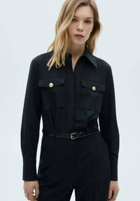 Svart skjorta med långa ärmar, knappstängning fram, skarp krage, två bröstfickor, guldknappar och en figursydd silhuett med ett svart bälte.