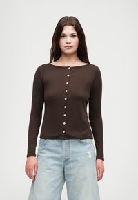 Haut marron à manches longues avec une texture côtelée et un large décolleté, présentant des boutons ressemblant à des perles sur le devant. Associé à un jean large bleu clair.