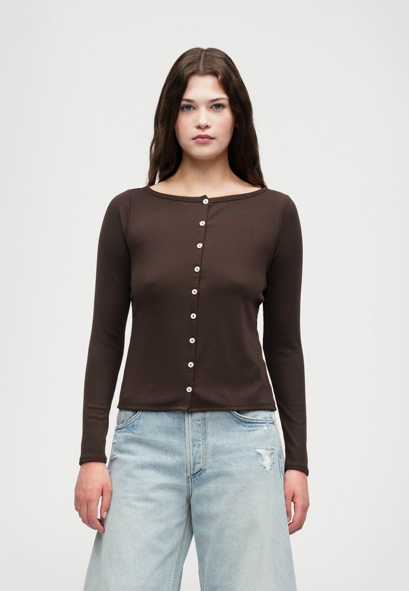 Haut marron à manches longues avec une texture côtelée et un large décolleté, présentant des boutons ressemblant à des perles sur le devant. Associé à un jean large bleu clair.