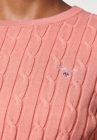 Pull en tricot rose avec un motif torsadé, doté d'un col rond et d'un petit patch logo en rouge, blanc et bleu. Texture douce.