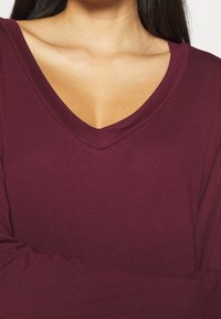 Bordeauxfarbene V-Ausschnitt-Bluse mit langen Ärmeln; weicher, elastischer Stoff; glatte Textur; moderater Ausschnitt; einfaches Design.