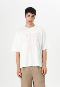 Bílý oversized tričko s potiskem "LOVERS IN" v světle růžové barvě. Krátké rukávy, kulatý výstřih, vyrobeno z měkké bavlny.