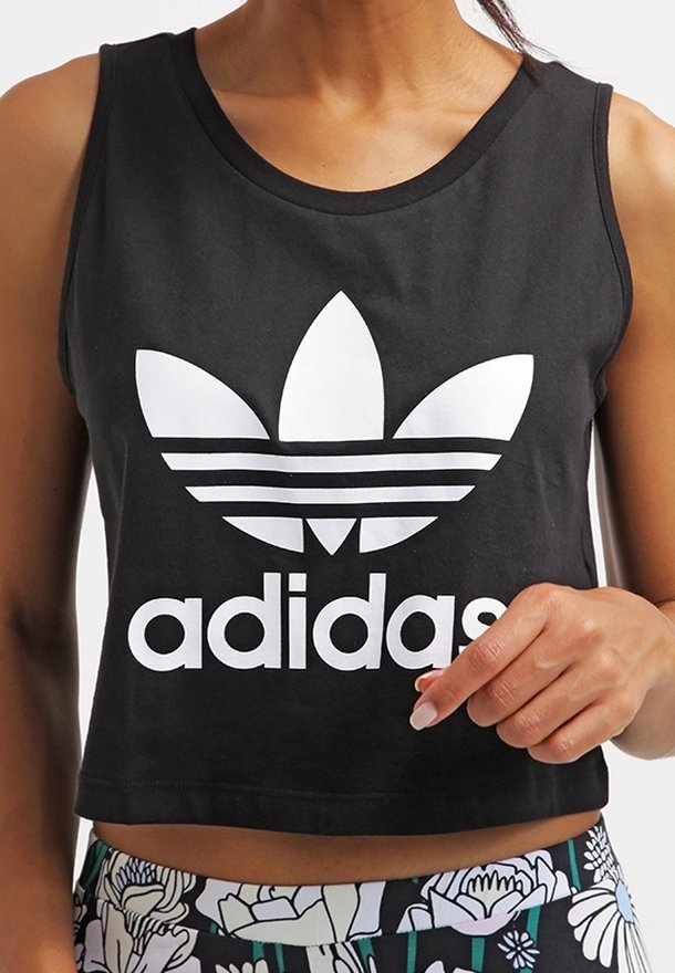 Kvinna som bär en svart ärmlös crop top med en stor vit Adidas-logga och byxor med blommönster.