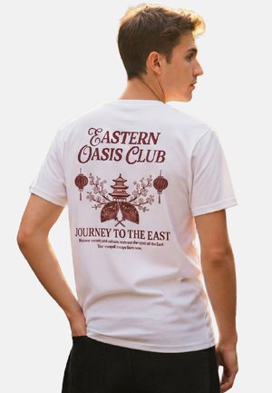 Young man facing sideways wearing white t-shirt with brown Asian-themed design and text "Eastern Oasis Club, Journey to the East."
