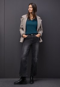 Cappotto di lana beige con collo oversize, top fitted teal, jeans neri a gamba larga e stivali neri alla caviglia. Texture morbide e colori neutri.