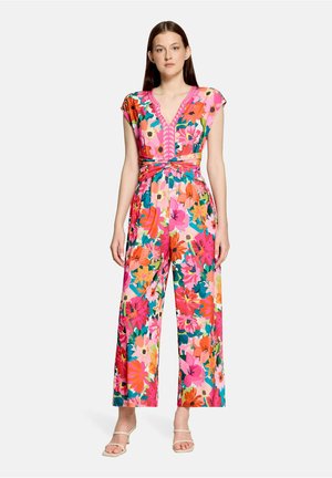 Kobieta stojąca, ubrana w kolorowy kwiatowy kombinezon z szerokimi nogawkami, dekoltem w serek i krótkimi rękawami, zestawiony z beżowymi szpilkami na paski.