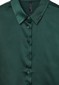 Camicia di raso verde scuro con colletto classico, sei bottoni e una texture liscia e lucida. Etichetta del marchio visibile sul collo.