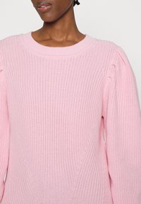 Maglione lavorato a maglia rosa chiaro con texture a coste, scollo rotondo ampio e maniche a sbuffo con dettagli sottili.
