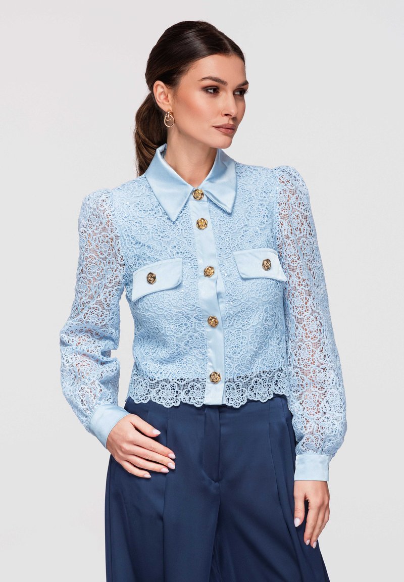 Femme portant un chemisier en dentelle bleu clair avec des boutons dorés et des poches sur la poitrine, associée à un pantalon taille haute bleu marine, debout avec une main sur la hanche.