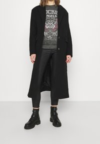 Person wearing a long black coat, graphic sweatshirt, black leather leggings, and black combat boots standing against a plain background.