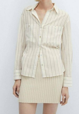 Kobieta nosząca kremową, pionowo paskowaną koszulę na guziki z dwoma kieszeniami na piersi oraz beżową, dzianą mini spódnicę.