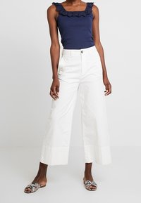 Marineblå tanktop med blonder, hvide bukser med vidde og mønstrede sandaler. Fokus på teksturer: blødt stof og blonderdetaljer.