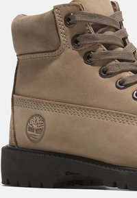 Stivaletto in suede beige con lacci marroni, occhielli in metallo e suola in gomma nera. Presenta un logo embossato e dettagli di cucitura a contrasto.