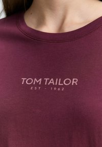 T-shirt bordeaux en coton doux, avec un col rond et un logo imprimé "TOM TAILOR EST - 1962" en couleur claire sur la poitrine.