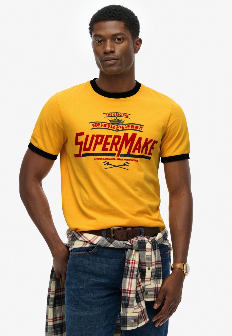 Yellow cotton t-shirt with black trim features bold red "SUPERMAKE" graphic. Worn with dark jeans and a plaid shirt tied around the waist.