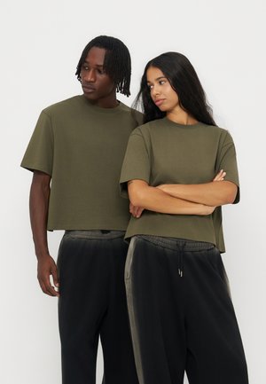 Zwei Personen tragen olivgrüne bauchfreie T-Shirts und schwarze Jogginghosen, stehen dicht nebeneinander und schauen seitlich vor einem schlichten Hintergrund.