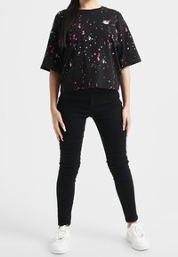 Mujer con camiseta negra de patrón salpicado, pantalones ajustados negros y zapatillas blancas de pie contra un fondo liso.