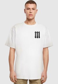 Balta kokvilnas T-krekls ar īsām piedurknēm, uz kura kreisajā krūtīs ir melns burts "M" logotips. Vienkāršs, brīvs piegriezums un apaļš kakla izgriezums.