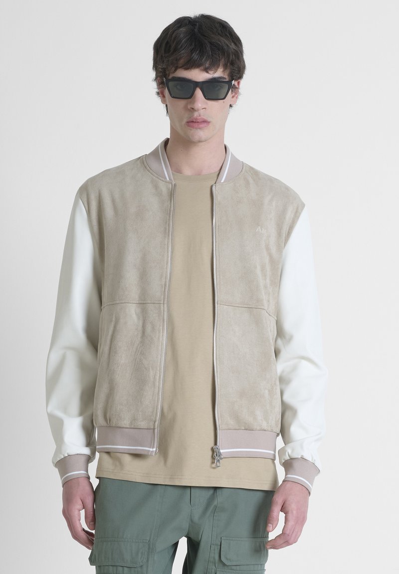 Chaqueta bomber de ante beige con mangas blancas, puños acanalados y cierre de cremallera. Lleva puesta una camisa color tostado y pantalones cargo verdes. Incluye gafas de sol.