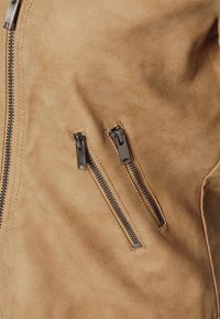 Wildlederjacke in Tan mit glatter Textur. Verfügt über zwei mit Reißverschluss versehene Brusttaschen mit silberner Reißverschluss-Hardware für sichere Aufbewahrung.