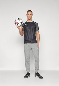 Fekete, rövid ujjú sportpóló, texturált mintával, szürke sportnadrág és fekete sportcipő, miközben egy focilabdát tart.