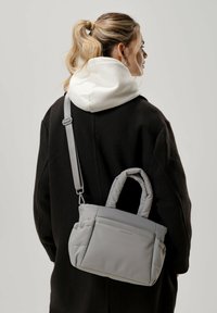 Szara torba typu puffer z podwójnymi uchwytami i regulowanym paskiem. Gładka faktura, prostokątny kształt i subtelny detal logo z boku.