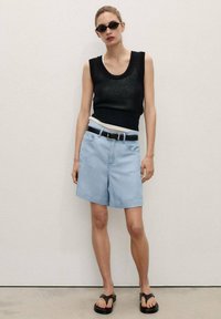 Haut sans manches noir, short en denim bleu clair, ceinture blanche et sandales noires. Les accessoires incluent des lunettes de soleil sombres. Tissu lisse et coupe décontractée.