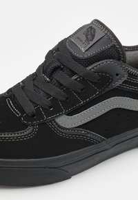 Gros plan d'une basket Vans noire avec une bande latérale grise, des lacets noirs, une languette texturée et des détails visibles de coutures blanches.