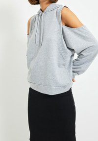 Šedá mikina s výřezy na ramenou, nastavitelnou kapucí se stahovací šňůrkou, volným střihem a žebrovaným lemem, spárovaná s černou sukní. Měkká textilie.
