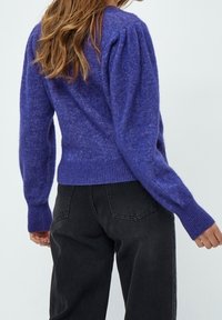 Woman wearing a purple long-sleeve fuzzy sweater with puffed shoulders and black high-waisted jeans, standing against a plain background.