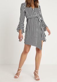 Schwarz-weiß gestreiftes Wickelkleid mit Glockenärmel, einem gebundenen Taillenbund und einem asymmetrischen Saum. Kombiniert mit hellen, schlichten Sandalen.