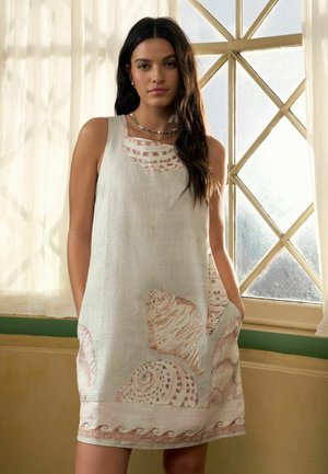 Woman with long dark hair wearing a sleeveless beige dress with seashell prints, standing in front of a window with white curtains.