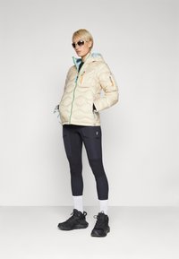 Beige gewatteerde jas met een capuchon, voorzien van een lichtblauwe voering, oranje en roze accenten, gecombineerd met zwarte leggings en stevige zwarte sneakers.