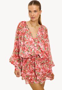 Vrouw in een roze, rode en groene bloemenjurk met lange puffmouwen, V-hals en gelaagd ruchezoom, geaccessoireerd met gouden hoepel oorbellen en een ketting.