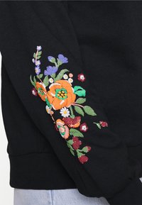 Manche en tissu noir avec des fleurs brodées colorées, comprenant des coquelicots orange, des marguerites violettes et blanches, et des feuilles vertes.