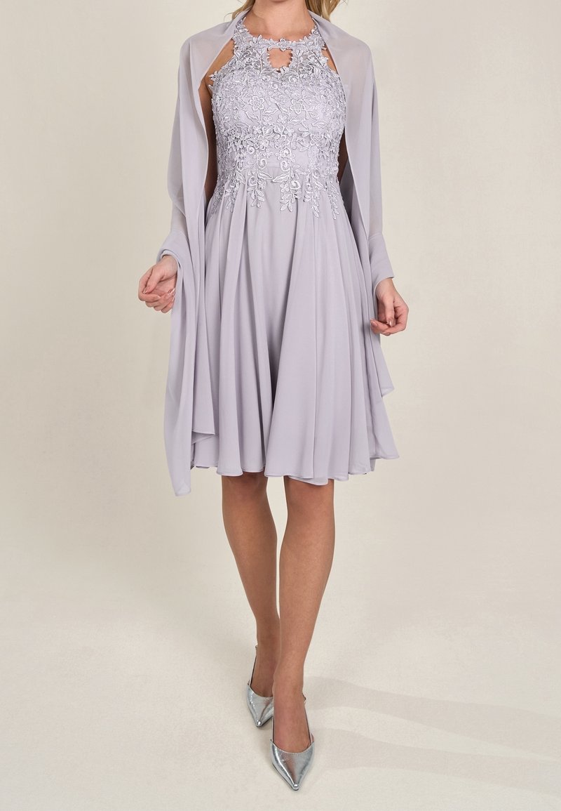 Femme portant une robe lavande sans manches avec un corsage en dentelle florale, un châle assorti et des escarpins argentés à bouts pointus, debout devant un fond uni.